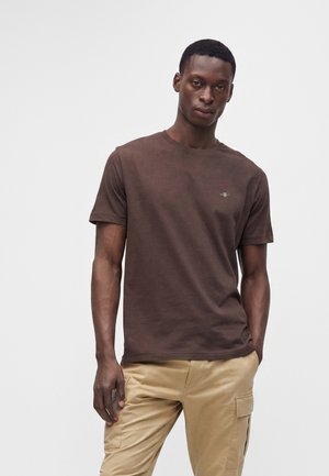 Homme debout portant un t-shirt marron à manches courtes et un pantalon cargo beige sur un fond blanc uni.