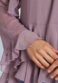 Blusa di un delicato viola chiaro con maniche a volant, realizzata in tessuto testurizzato. I polsini presentano cuciture raffinate e dettagli a volant sovrapposti.