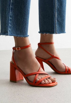 Des pieds portant des sandales à talons blocs orange vif à brides, associées à un jean bleu court sur un sol clair.