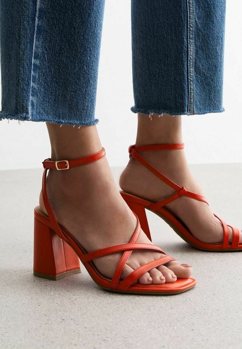 Des pieds portant des sandales à talons blocs orange vif à brides, associées à un jean bleu court sur un sol clair.