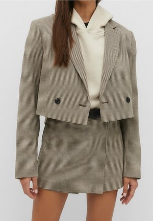 Femme portant un blazer court à motif pied-de-poule marron et une jupe assortie avec un sweat à capuche beige en dessous, posant devant un fond uni.
