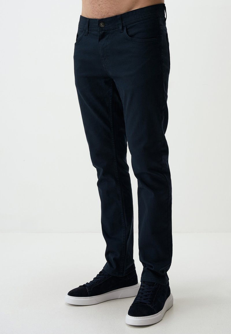 Ciemnoniebieskie dżinsy slim fit z bawełnianej tkaniny o gładkiej fakturze, z przednimi kieszeniami i standardowym zapięciem na guzik. Połączone z czarnymi sneakersami.