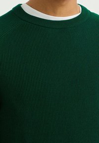 Groene ribgebreide trui met een ronde halslijn, gelaagd met een witte rondhals t-shirt eronder. De textuur lijkt zachter en comfortabel aan te voelen.