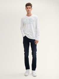 Camisa blanca de manga larga con texto turquesa, jeans oscuros y zapatillas blancas. Diseño sencillo, corte relajado y textura suave.