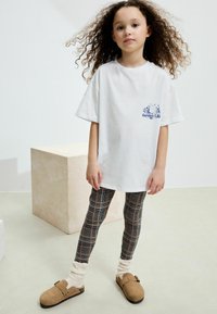 Vit bomulls-T-shirt med trycket "Garden Life", kombinerad med rutiga leggings i blått, orange och svart, samt bruna mockaklackar med spänne.