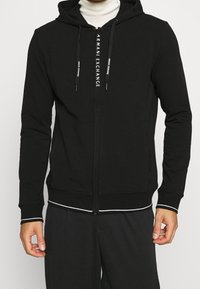 Felpa nera con zip e testo "Armani Exchange" sui laccetti e sulla zip, indossata sopra un dolcevita bianco e pantaloni neri coordinati.