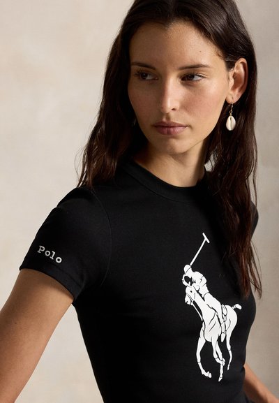 Γυναίκα με μακριά σκούρα μαλλιά, φορώντας μαύρο πουκάμισο πόλο με λευκό λογότυπο παίκτη πόλο και σκουλαρίκια σε σχήμα κοχυλιού, κοιτάζοντας προς τα δεξιά της.