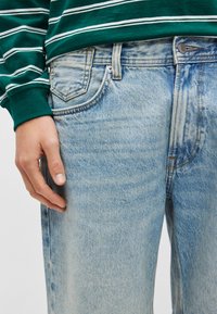 Ανοιχτό μπλε τζιν παντελόνι με ξεβαμμένο φινίρισμα, διαθέτει πέντε τσέπες και διακριτική αντίθεση στο ράψιμο. Το χέρι ακουμπά στην πλαϊνή τσέπη.