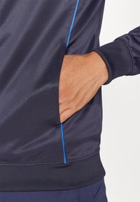 Marine athletisk genser i glatt, lett stoff med en sidelomme og en kontrast blå detalj på sømmen.