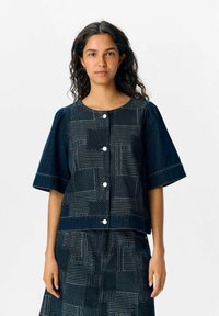 Marinblus i denim med puffärmar, rund halsringning och knappstängning. Strukturerat mönster med linjer och prickar över tyget.