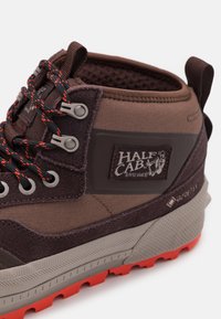 Brun vandringsko med en blandning av tyg och gummi, med grå öglor, orange snören och en "HALF CAB"-logotyp. Mönstrad sula.