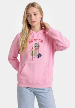 Ung kvinde iført en lyserød hættetrøje med en kattegrafik og teksten "Happy Cat", kombineret med lyseblå jeans og blå øreringe i hoop-design.