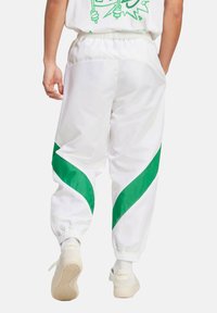 Bijele trenirke s zelenim dijagonalnim prugama, elastičnim pojasom i manžetama, izrađene od glatkog, sjajnog materijala.