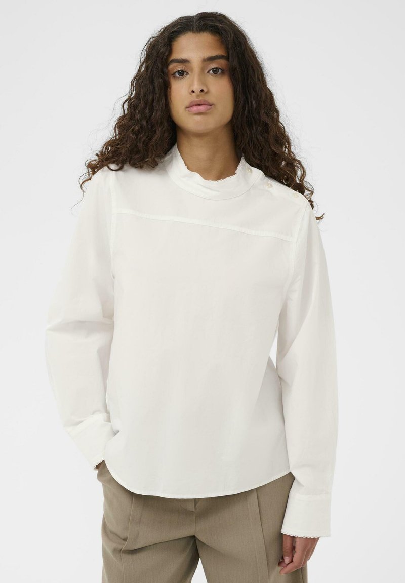 Biała bluzka z długim rękawem i wysokim kołnierzem, z delikatnym falbanym wykończeniem, poziomą szwem oraz akcentami w postaci guzików perłowych na jednym ramieniu.