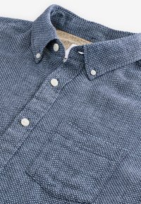 Plava košulja sa uzorkom od teksturiranog tkanja, s okovratnikom na kopčanje, bijelim gumbima i džepom na lijevoj strani prsa.