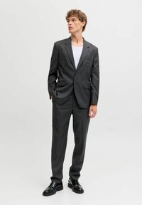 Ciemny garnitur w prążki z dopasowaną marynarką i spodniami, wykonany z gładkiej tkaniny; zestawiony z białym podkoszulkiem z okrągłym dekoltem i czarnymi butami.