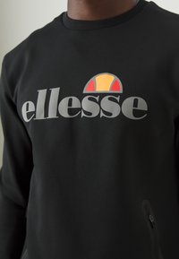 Felpa nera con il logo grigio "ellesse" e un emblema a semicircolo arancione e rosso sul petto.