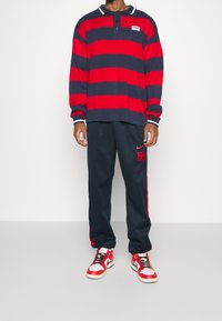 Camisa de manga larga a rayas rojas y marinas con cuello y botones, combinada con pantalones de chándal marinos con rayas rojas en los laterales y un parche de logo. Zapatillas rojas y blancas.