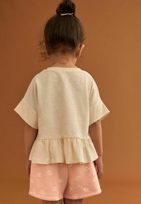 Ljusbeige kortärmad topp med peplum-nertill och rosa shorts med blommönster. Bakgrunden är en enfärgad brun färg.