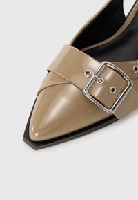 Beige patent leather pointed-toe slingback shoe with a wide strap and a silver buckle detail, featuring a contrasting black sole.