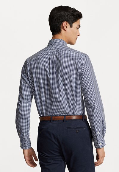 Chemise de costume à carreaux bleus et blancs pour homme, à manches longues, avec des poignets à bouton et un pli au dos, portée avec un pantalon sombre et une ceinture marron.