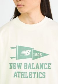 Camiseta color crema con un gran gráfico en verde menta de un estandarte que cuenta con el texto "NB 1906" y "NEW BALANCE ATHLETICS" en letras grandes.