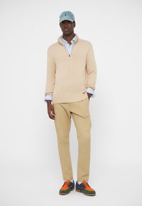 Beige gebreide sweater met een ritssluiting, lichtblauw gestreept overhemd eronder, beige cargobroek en multicolour sneakers; blauwe pet.