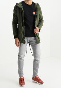 Chaqueta ligera verde con capucha sobre un sudadera negra con logo, jeans grises y zapatillas naranjas con detalles en blanco. Atuendo urbano casual.