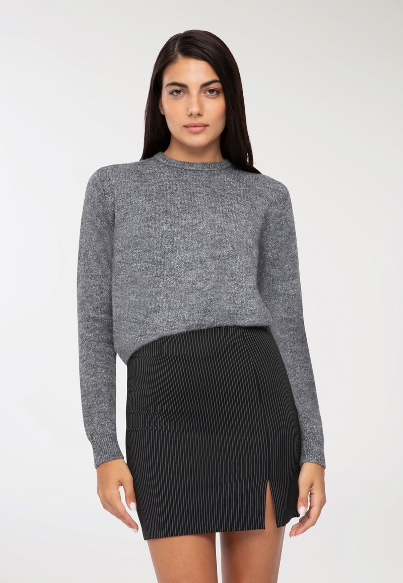 Una donna con i capelli lunghi e scuri indossa un maglione grigio a maglia e una gonna nera a righe con apertura frontale, in piedi davanti a uno sfondo bianco semplice.
