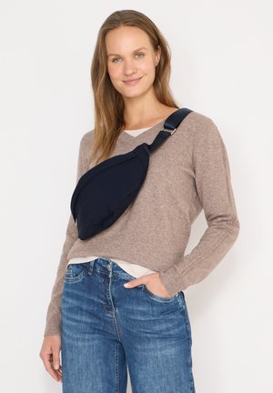 Femme souriante aux longs cheveux, portant un pull beige, un jean bleu et un sac bandoulière bleu marine, sur un fond clair uni.