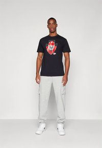 Černé tričko s grafikou červené fotbalové helmy, doplněné šedými cargo tepláky a bílými teniskami. Jednoduchý design a volný střih.