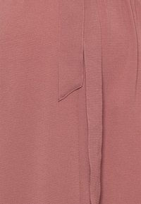 Różowy strój z teksturowanej, miękkiej tkaniny. Posiada akcent w postaci związanej tasiemki oraz luźny krój. Widoczny detal przeszycia wzdłuż krawędzi.