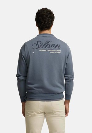 Hombre con cabello rizado que lleva una sudadera azul Silbon y pantalones beige, de espaldas, mostrando el logo y texto en la parte trasera de la sudadera.