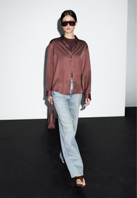 Brown satin button-up shirt, light blue straight-leg jeans, brown sandals, and small maroon handbag; model wears dark sunglasses.