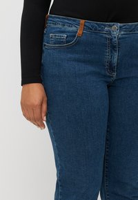 Blue denim jeans featuring a mid-rise waist, curved pocket seams, and a contrast brown leather accent on the waistband. Smooth texture.