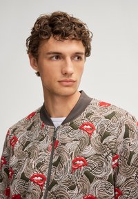 Giubbotto bomber con base khaki, tessuto testurizzato e un motivo vorticoso con labbra rosse e accenti rosa. Collo grigio e zip.
