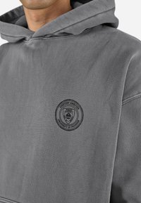 Grijze hoodie van zacht materiaal met een opvallende zwarte geborduurde patch. Ontwerp omvat een ruime capuchon en een relaxte pasvorm.
