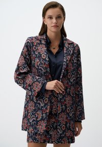 Blazer à motif floral bleu marine, rouge et vert, avec des manches longues et des revers à crans, associé à une chemise bleu foncé.