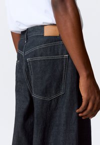 Mørke denimjeans med rett snitt, med kontraststing, to baklommer og et brunt lærmerke på linningen.