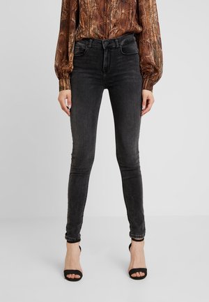 Jeans negros deslavados de corte ajustado combinados con blusa marrón con estampado de serpiente y tacones abiertos negros, en una persona de pie sobre un fondo liso.