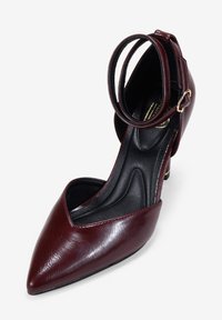 Zapato de tacón burdeos con punta afilada, correa en el tobillo y textura de cuero suave. Plantilla negra con contorno para soporte.