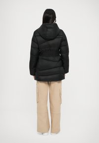 Svart puffjakke med hette, innsnevret midje og horisontalt quiltet mønster, kombinert med beige cargo-bukser og hvite sko.