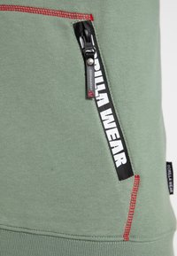 Sudadera verde con un tejido texturizado, un bolsillo con cremallera que lleva una etiqueta en blanco y negro de "GORILLA WEAR", y detalles de costura en rojo contrastante.