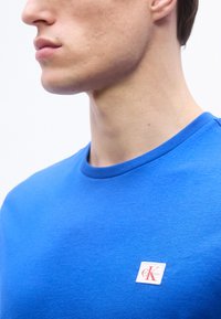 Νεαρός άνδρας που φοράει μπλούζα σε έντονο γαλάζιο χρώμα με στρογγυλή λαιμόκοψη και μικρό κόκκινο και λευκό λογότυπο Calvin Klein στο στήθος, με λευκό φόντο.