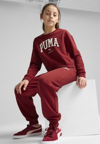 Sudadera maroon de PUMA con logo, pantalones de chándal maroon a juego y zapatillas rojas con detalles blancos, sobre un fondo neutro.