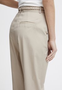 Beige skräddarsydda byxor med en slät textur, försedda med ett flätat bälte och en diskret bakficka. Enkelt och rent designlinjer.