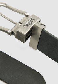 Cinturón de cuero negro con un broche de metal plateado que presenta un logotipo, hebilla rectangular y superficie texturizada. Bordes lisos y diseño minimalista.