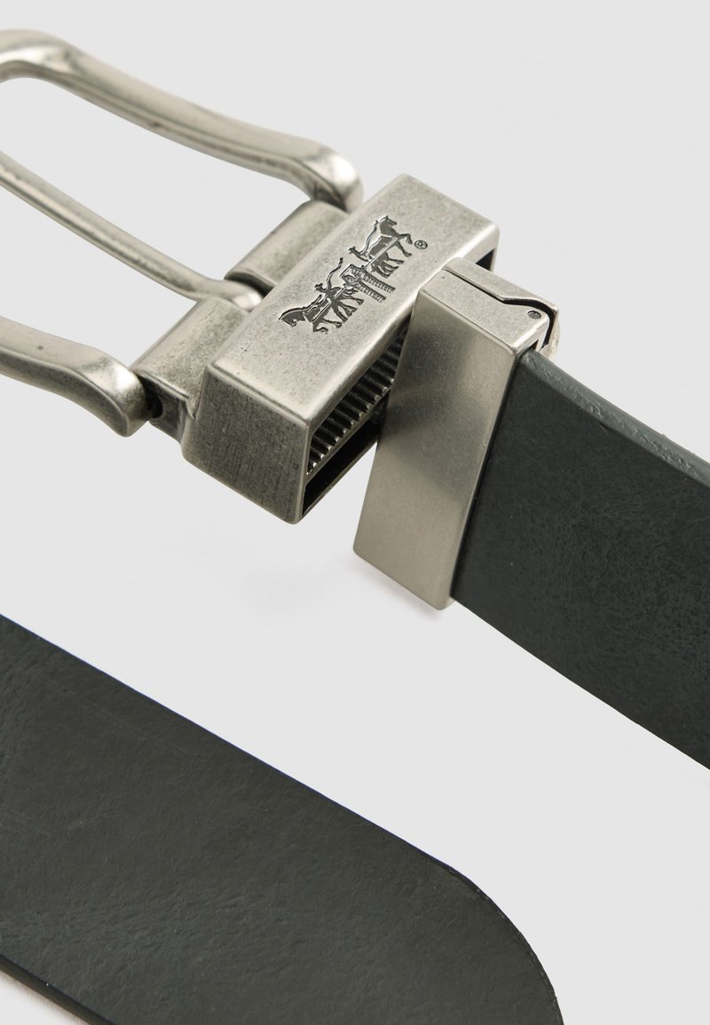 Sort læderbælte med en sølvmetalspænde, der har et logo, rektangulært spænde og tekstureret overflade. Glatte kanter og minimalistisk design.