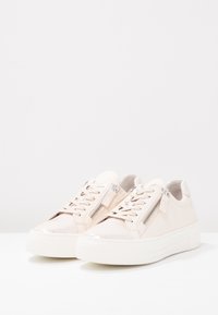 Baskets beiges avec un bout en cuir verni brillant, une tige texturée, une semelle blanche plate, et une fermeture éclair latérale pour un accès facile.