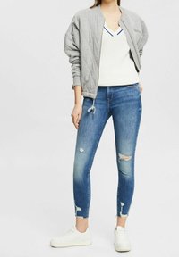 Grå vadderad bomberjacka över en vit stickad topp, i kombination med blå skinny jeans med slitna detaljer och fransade fållar, komplett med vita sneakers.